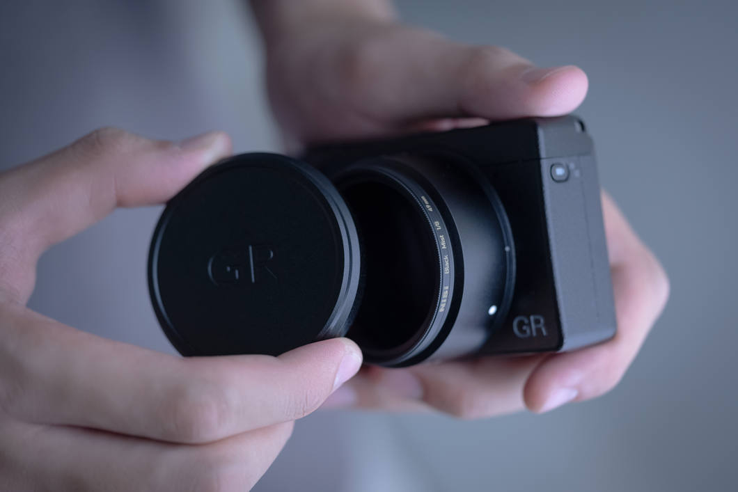 Ricoh GR IV i två händer med NiSis objektivlock med GR-logga på objektivet